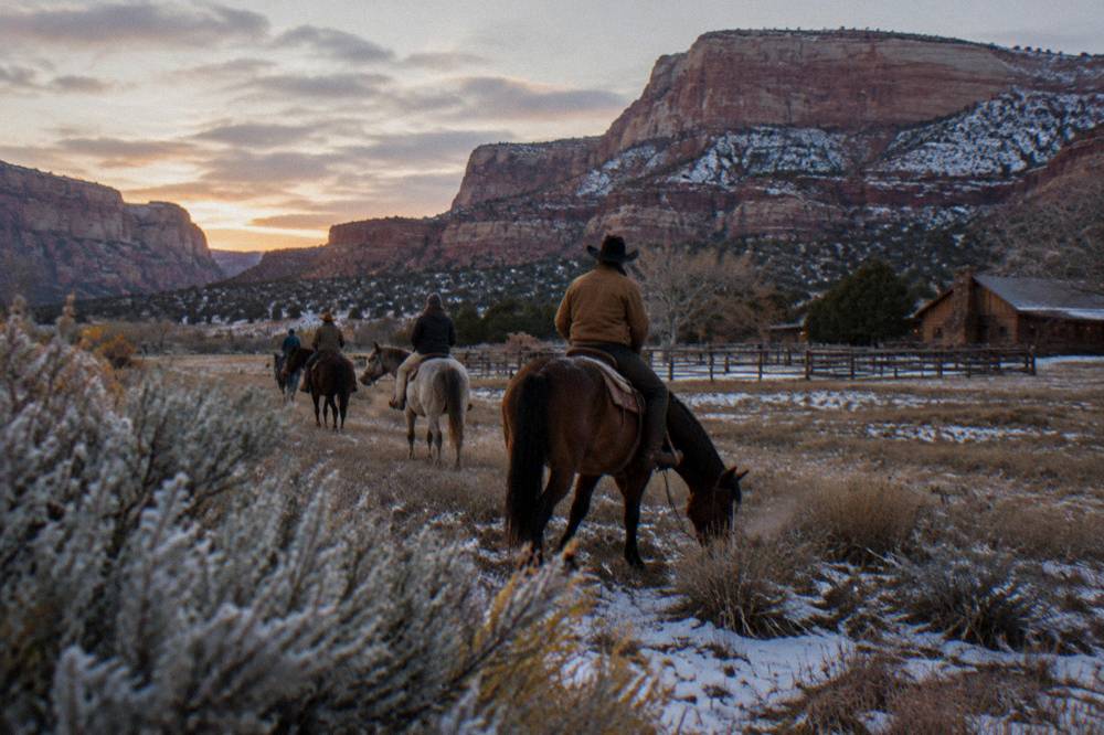 Horseback riders crossing open sage rangeland toward wide stratified cliffs at dusk